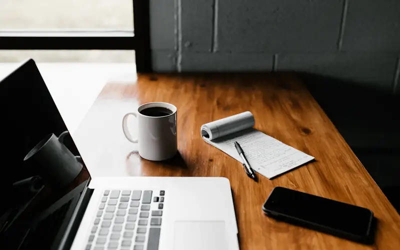 A laptop, a coffee and a notebook on a desk in the morning