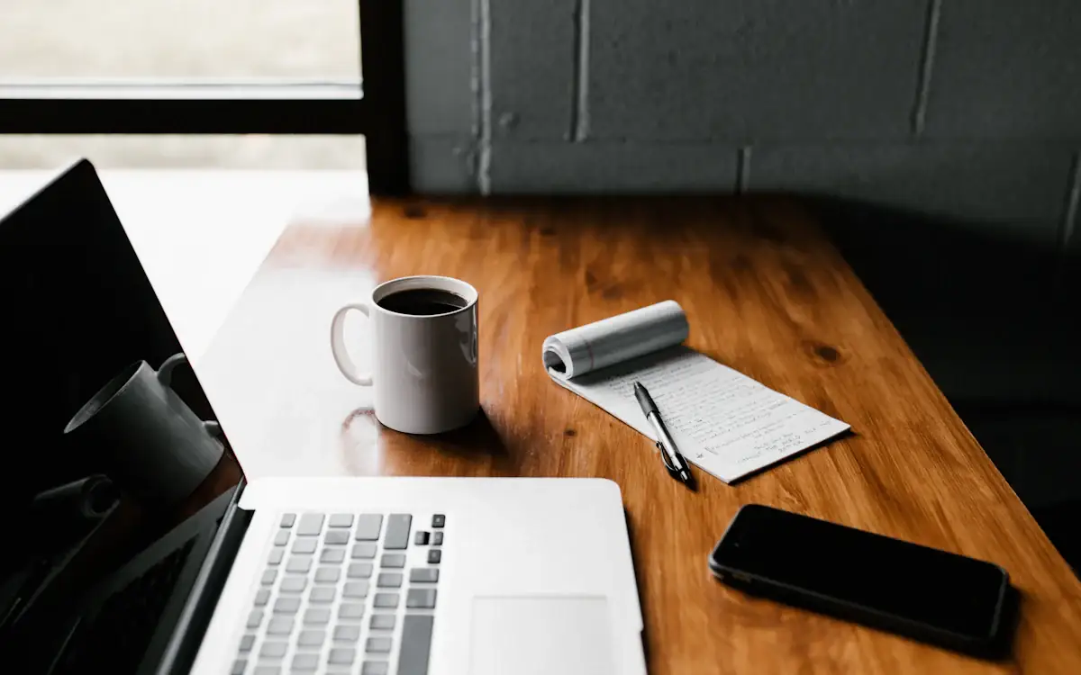 A laptop, a coffee and a notebook on a desk in the morning